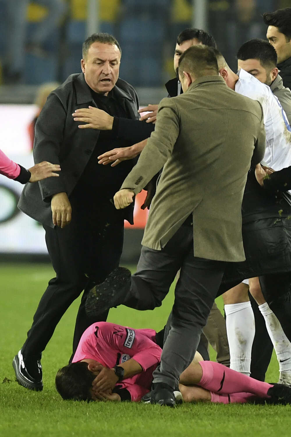 Neidentifikovaný muž kope tureckého arbitra Halila Umuta Melera (uprostred), ktorý leží na trávniku.